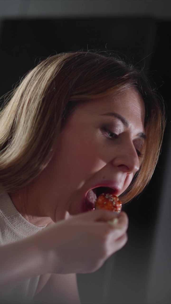 Primer plano de una mujer comiendo, centrándose en los movimientos de su boca y manos mientras da bocados. La escena se desarrolla sobre un fondo oscuro con iluminación suave que resalta las acciones del sujeto.