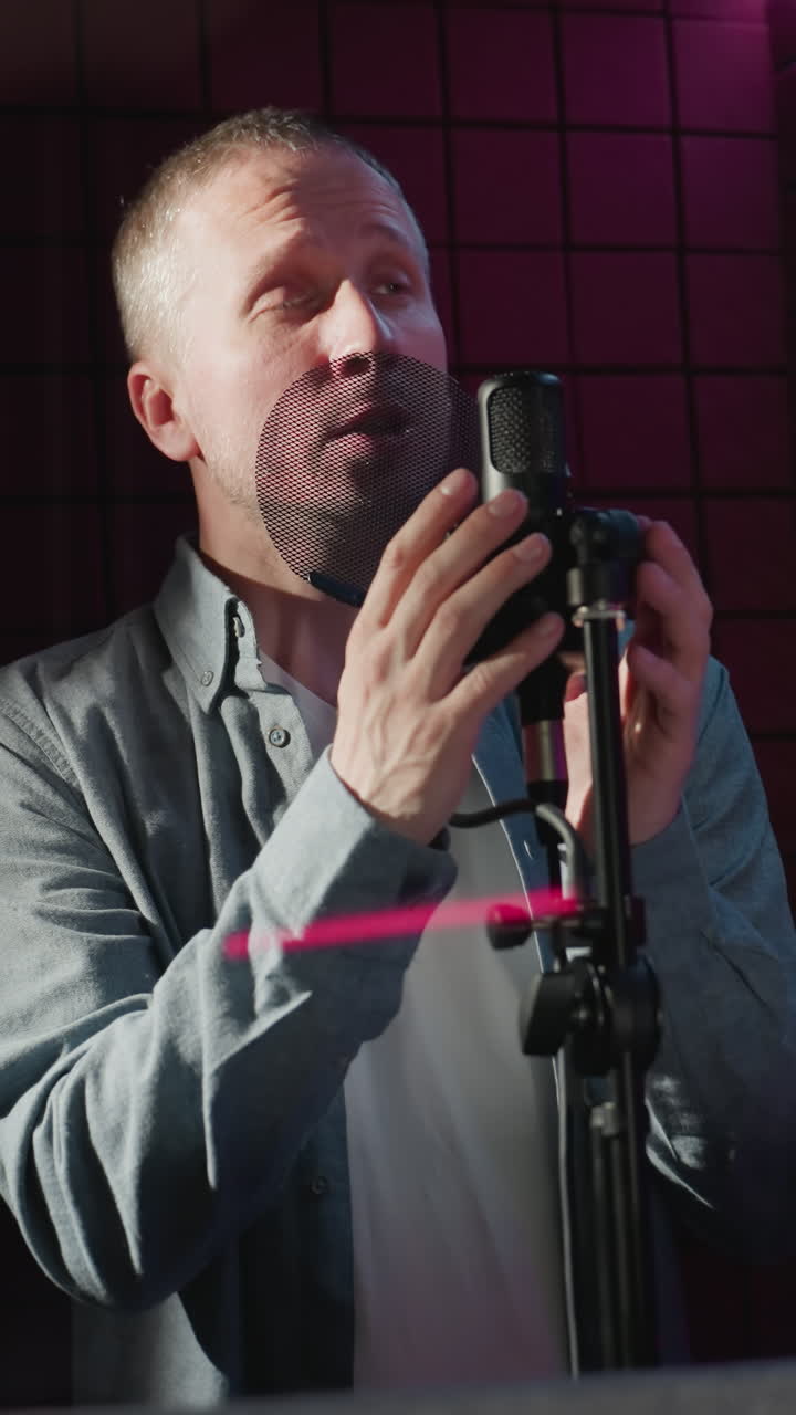 un hombre con una camisa de manga larga azul y camisa interior blanca canta apasionadamente en un micrófono dentro de un estudio a prueba de sonido rojo, la iluminación proyecta sombras dramáticas y mejora la atmósfera