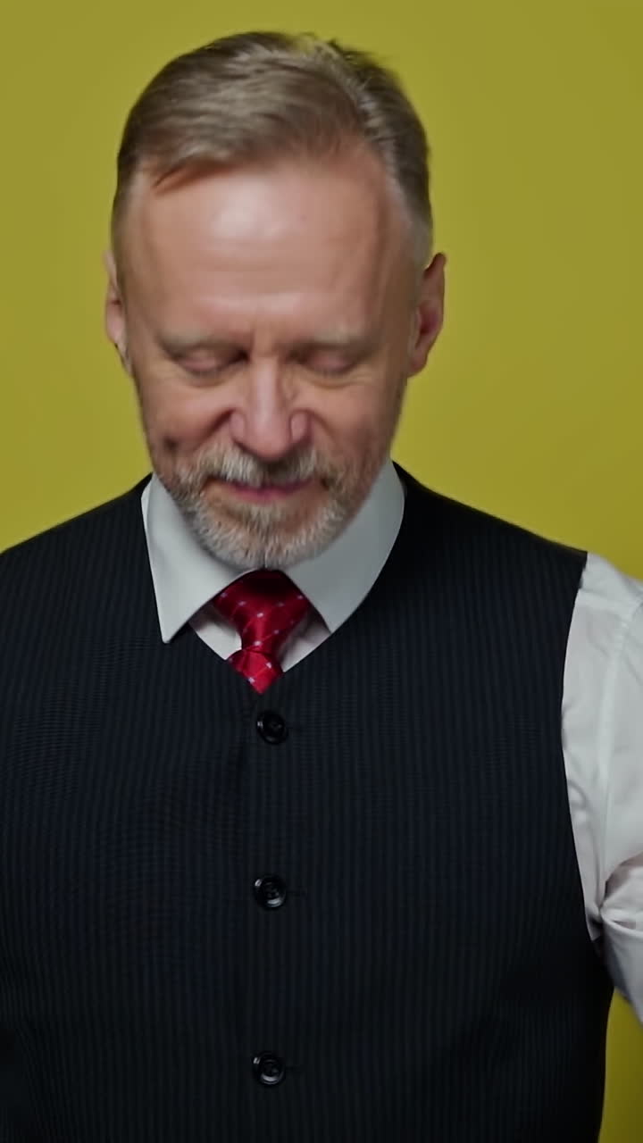 Portrait of a handsome grey-haired man. Mature man taking off his eyeglasses with a smile and looking at camera on yellow background. Vertical video