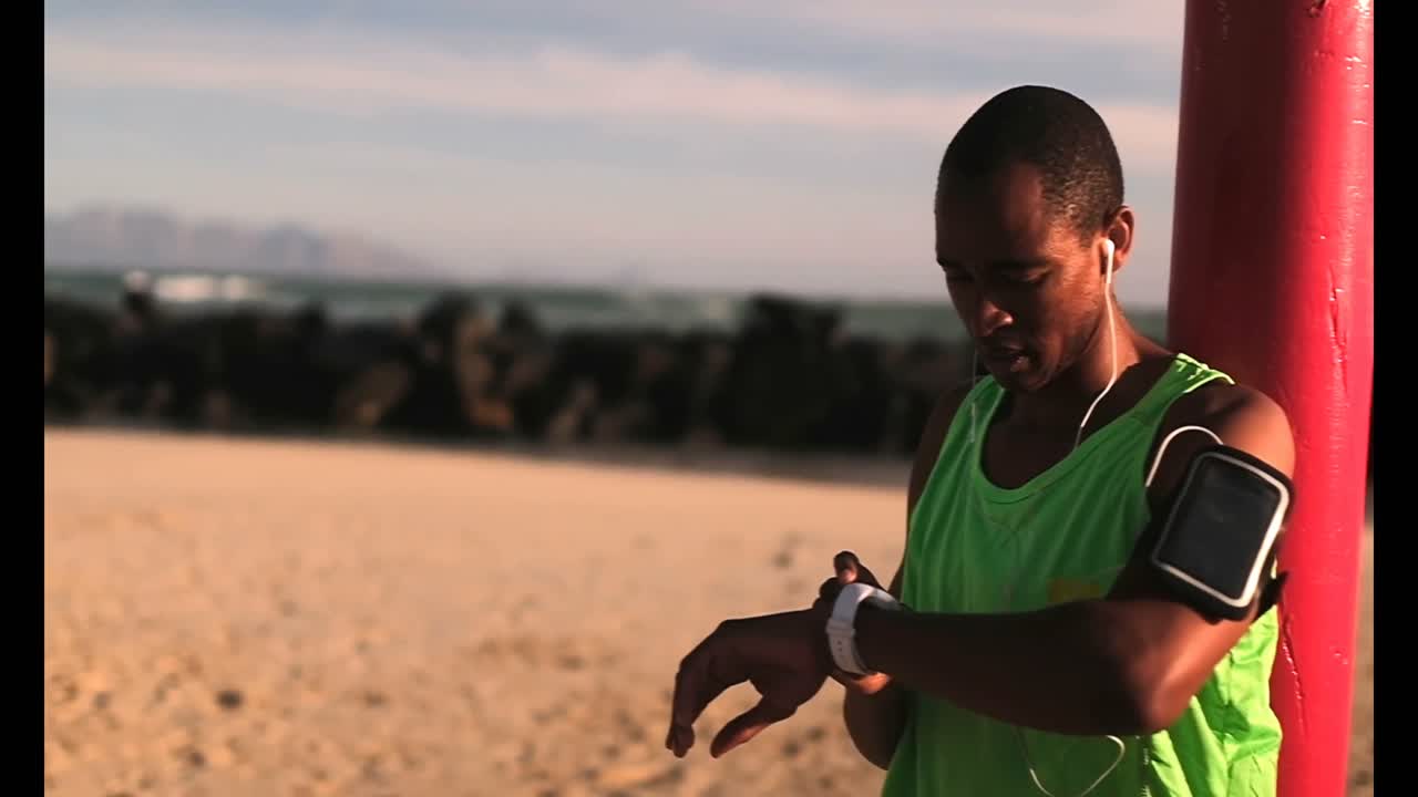 corredor afroamericano usando un reloj inteligente en la playa bajo el sol 4k