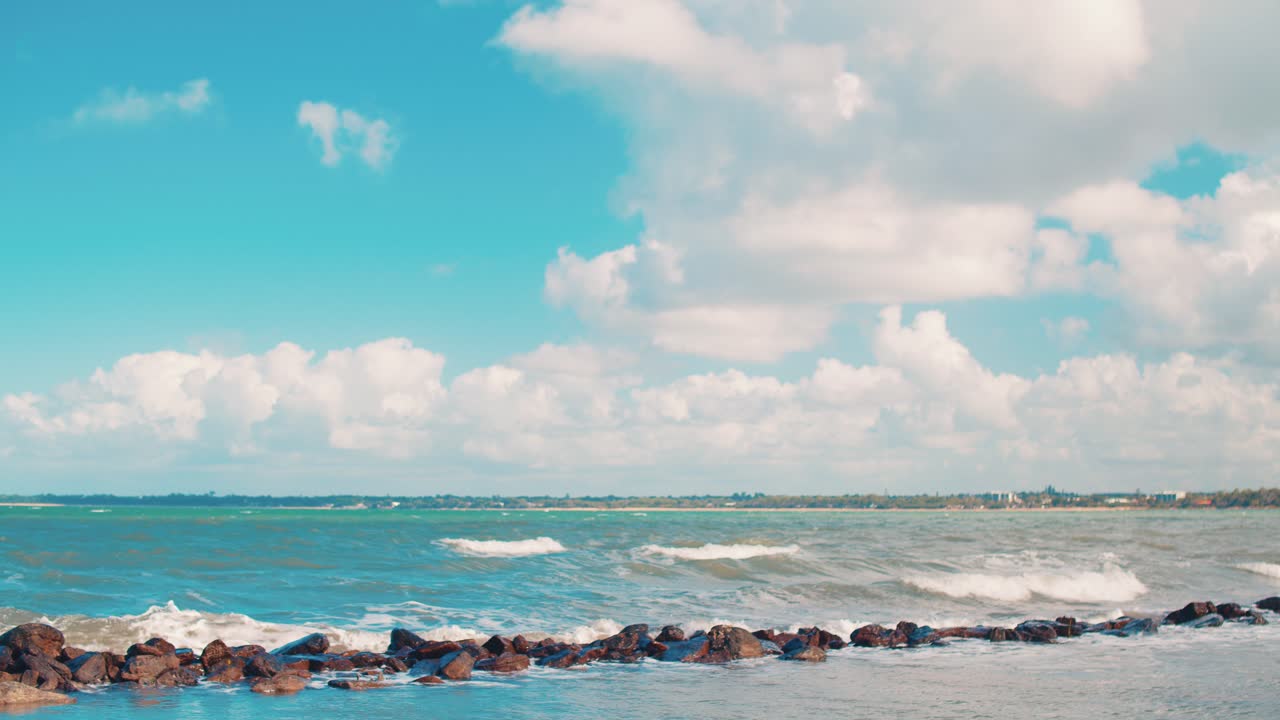 작은 파도가 아름다운 열대 푸른 바다에서 온전히 모양과 크기가 다른 바위들로 만들어진 짧은 벽에 부히며, 푸른 빛 하늘 아래, 색 구름이 떠다니고 있습니다.