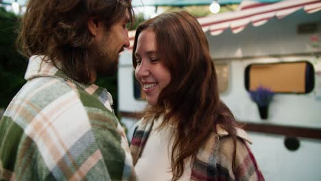 over the shoulder, a happy brunette girl in a pink checkered shirt hugs her brunette boyfriend in a green checkered shirt and kisses her on the forehead during her vacation and picnic outside the city in a camp in the summer