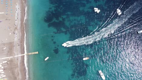 vista aérea de arriba hacia abajo de un barco a través de una hermosa agua azul turquesa llegando a positano en un soleado día de verano con una playa de arena blanca prístina