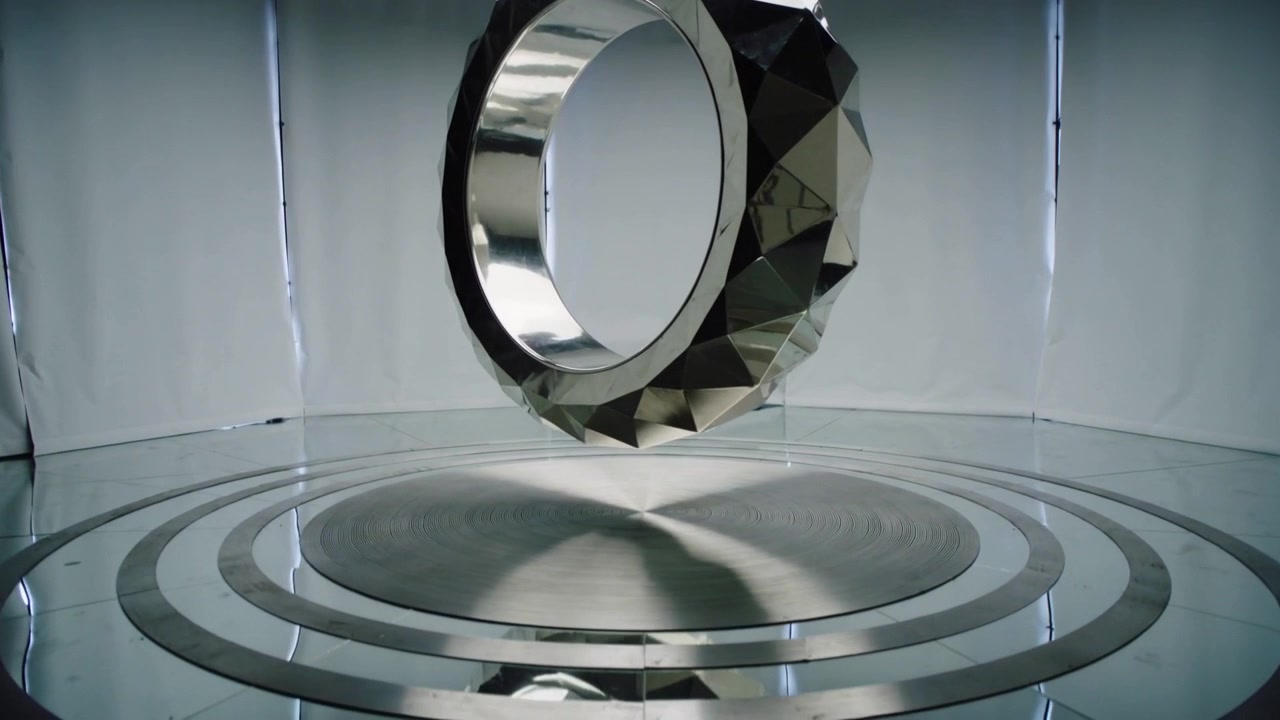 Motor rig lowering faceted metal torus on thin cable, rotating, settling onto center disc in studio