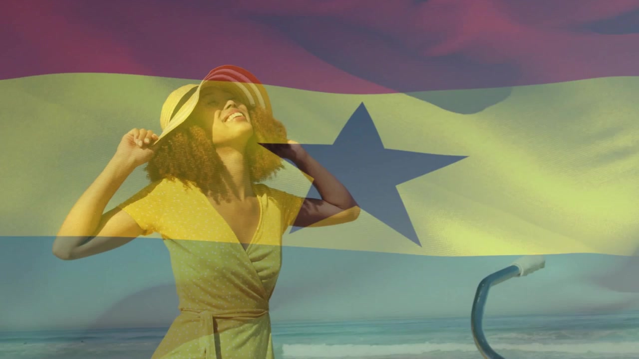 Frau am Strand, die im Wind ihren Hut zurechtrückt, den Kopf neigt, um die Augen zu beschatten, mit einer dünnen, wehenden Flagge im Bild, Reise.