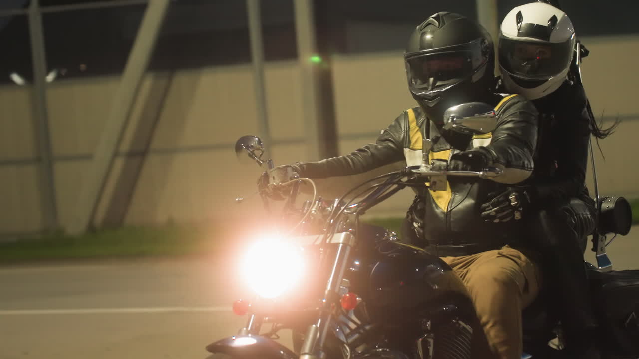 Two motorcyclists wearing helmets and leather jackets ride at night with bright headlight shining, moving along road beside tall fence under dark evening sky