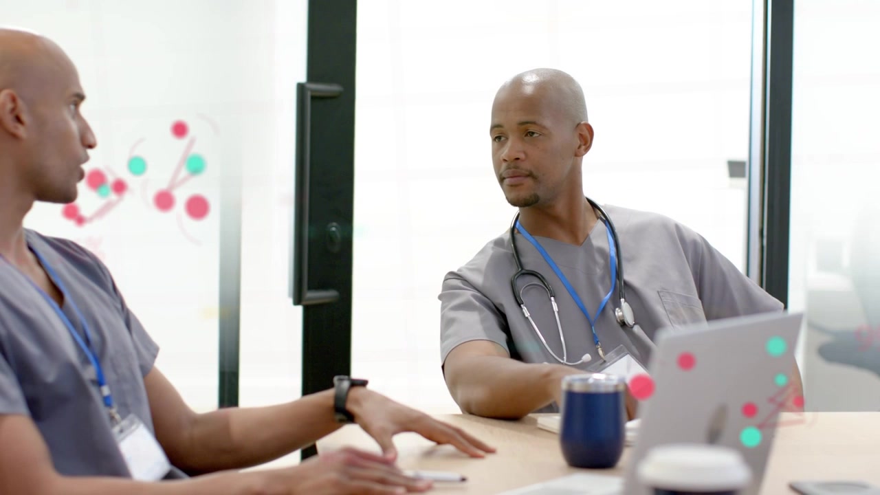 Médecin de gauche entrant et s'asseyant, entamant une discussion pendant qu'un collègue vérifie un ordinateur portable pour des données cliniques sur un écran en verre.