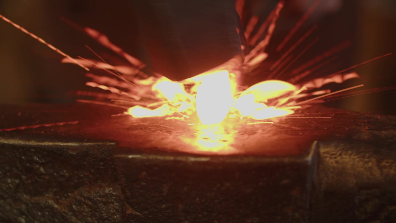 Primo piano di una punta di metallo incandescente martellata ripetutamente su un'incudine, con scintille che volano a ogni colpo, per mostrare l'arte del fabbro, l'artigianato e l'azione drammatica della fucina