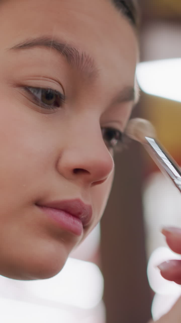 primer plano de una joven maquillándose con sombra de ojos en el backstage bajo luces brillantes; un estilista usa una brocha suave para difuminar, con movimientos rápidos y seguros, expresión concentrada, ambiente animado entre bastidores, reflejos en el espejo