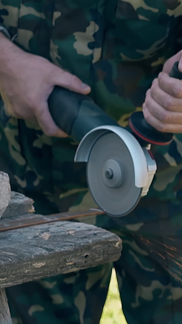 slow motion skilled worker in uniform cuts metal sheet with electrical tool against green grass on day