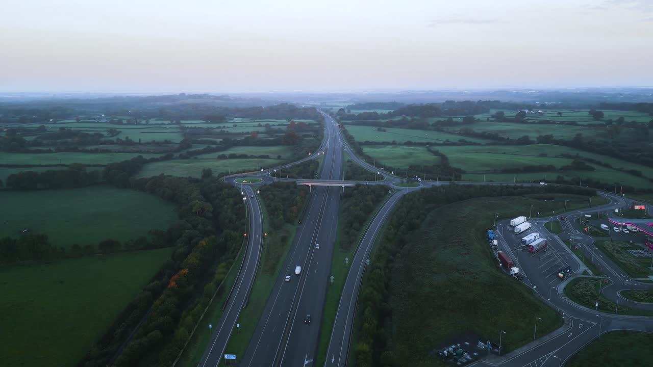 Circular drone footage of the M4 motorway interchange in Ireland. Highlights modern road infrastructure, roundabouts, and a service park amidst vast, lush green farmlands