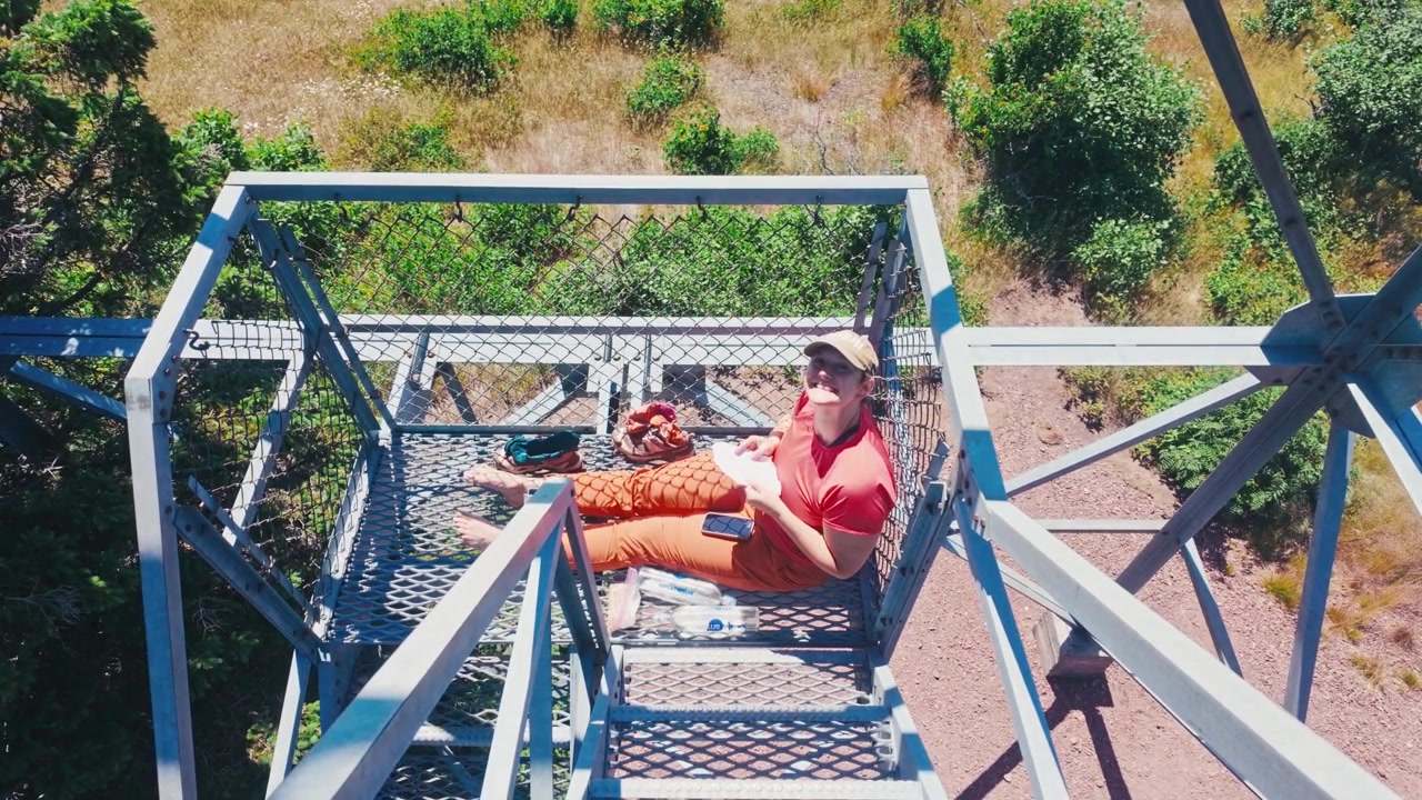 Un mochilero descansando en una escalera metálica de una torre de vigilancia y sonriendo a la cámara con un telón de fondo de naturaleza salvaje