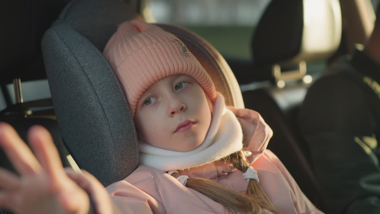 핑크색 뜨개질 모자와 색 스카프를 입은 어린 소녀가 차 안에서 조용히 앉아서 손을 부드럽게 흔들고 있다.