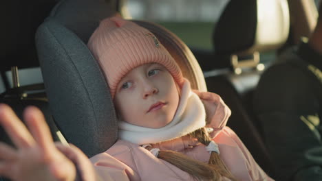 a young girl, dressed in a pink knit hat and white scarf, sits peacefully in a car, gently waving her hand. a man is seated beside her