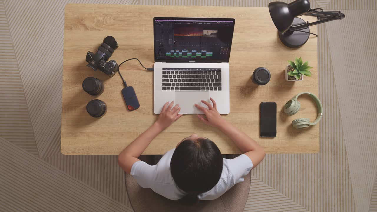 Top View Of Asian Woman Editor With Headphone Having A Backache While Sitting In The Workspace Using A Laptop Next To The Camera Editing The Video At Home