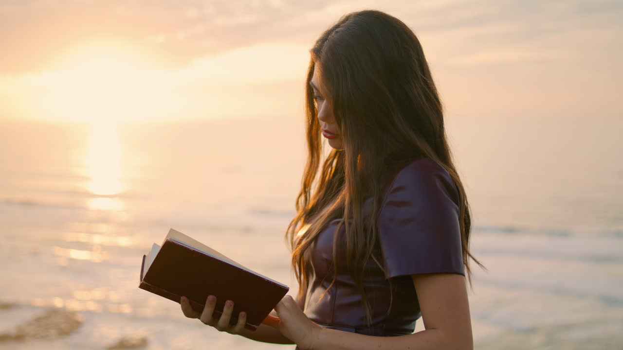 Garota lendo livro ao pôr do sol em close-up. Morena segurando livro de pé na beira-mar.