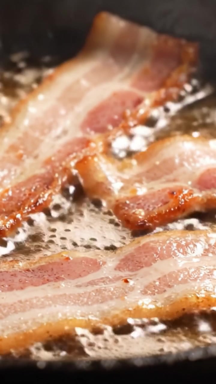 Sydende bacon steger til perfektion: Et nærbillede af sprøde strimler, der bruner på en varm pande, afgiver lækre aromaer og lyde af stegende fedt.