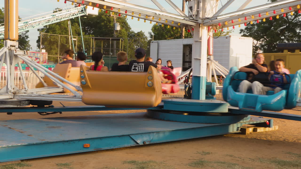 A slow-motion shot captures a carnival ride spinning riders around, with the sun shining brightly, creating a lively and exciting atmosphere.
