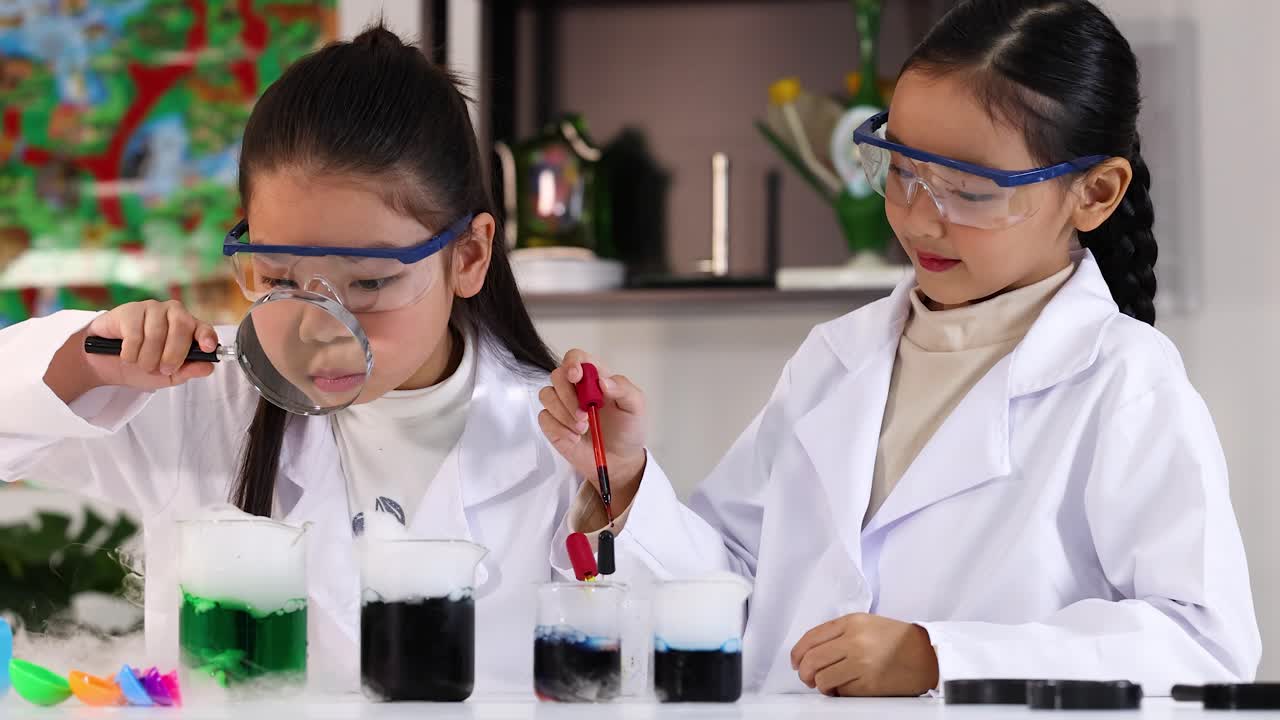 Two young Asian girls wear lab coats and safety goggles while conducting colorful chemistry experiments