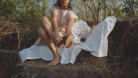 Slow-motion-handheld-panning-shot-of-a-beautiful-indian-model-sitting-on-a-white-sheet-in-the-garden-taking-off-her-shoe-on-a-sunny-day