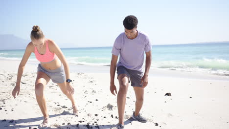 una joven caucásica y un hombre biracial están listos para correr en una playa soleada.