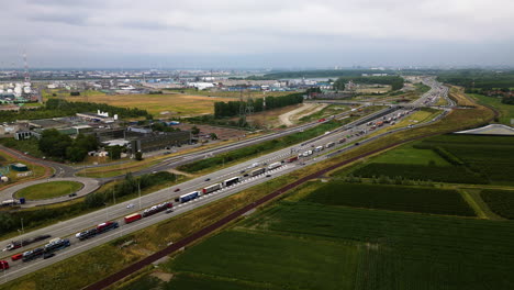 LKW-Verkehr-Auf-Der-Autobahn-In-Der-Nähe-Von-Landwirtschaftlichen-Feldern-Und-Industriegebieten,-Luftaufnahme