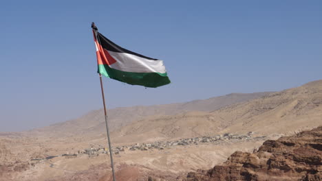 Flagge-Jordaniens,-Die-An-Sonnigen-Tagen-über-Trockener-Felsiger-Berglandschaft-Mit-Klarem-Himmel-Weht,-Mit-Der-Modernen-Petra-Im-Hintergrund