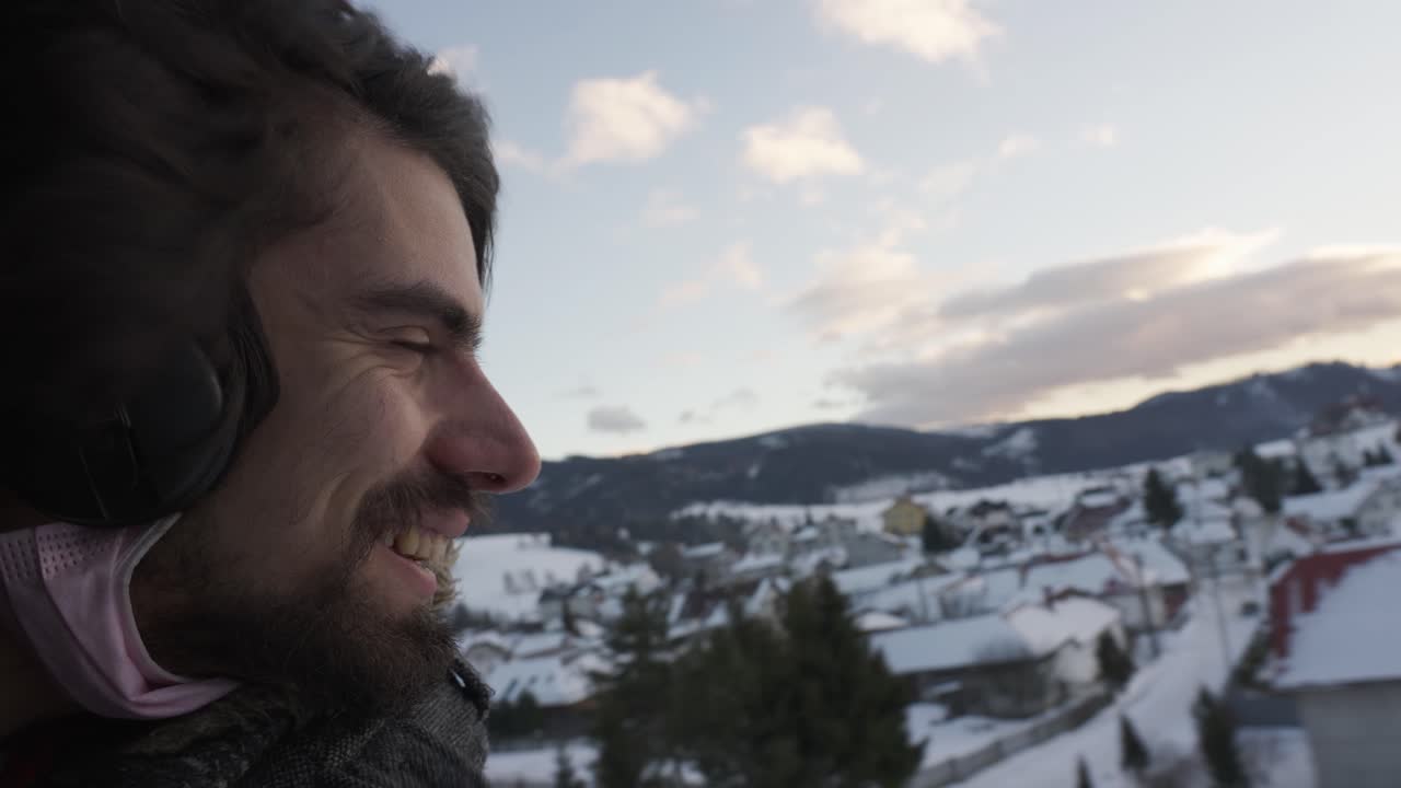 Uomo barbuto con le cuffie che sorride con la testa fuori dal finestrino aperto sul treno in un paesaggio invernale innevato, Slovacchia.
