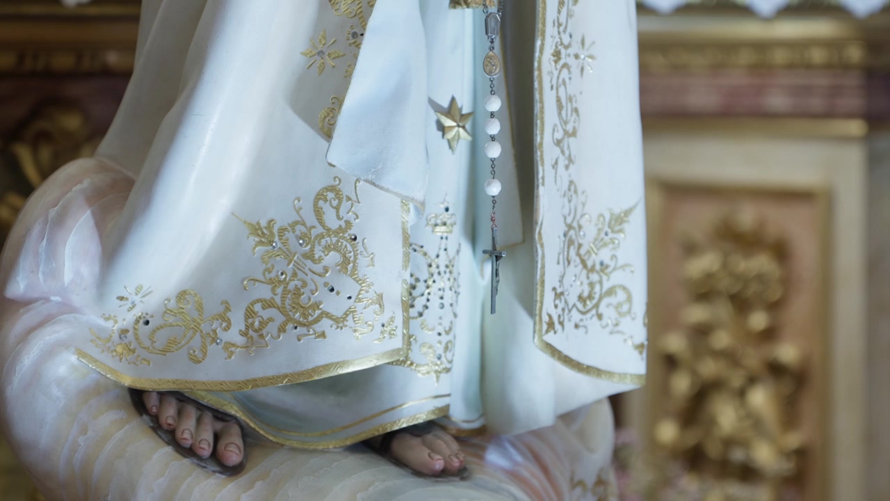 Dettaglio della statua di Nostra Signora di Fatima con rosario, veste ricamata in oro e piedi.