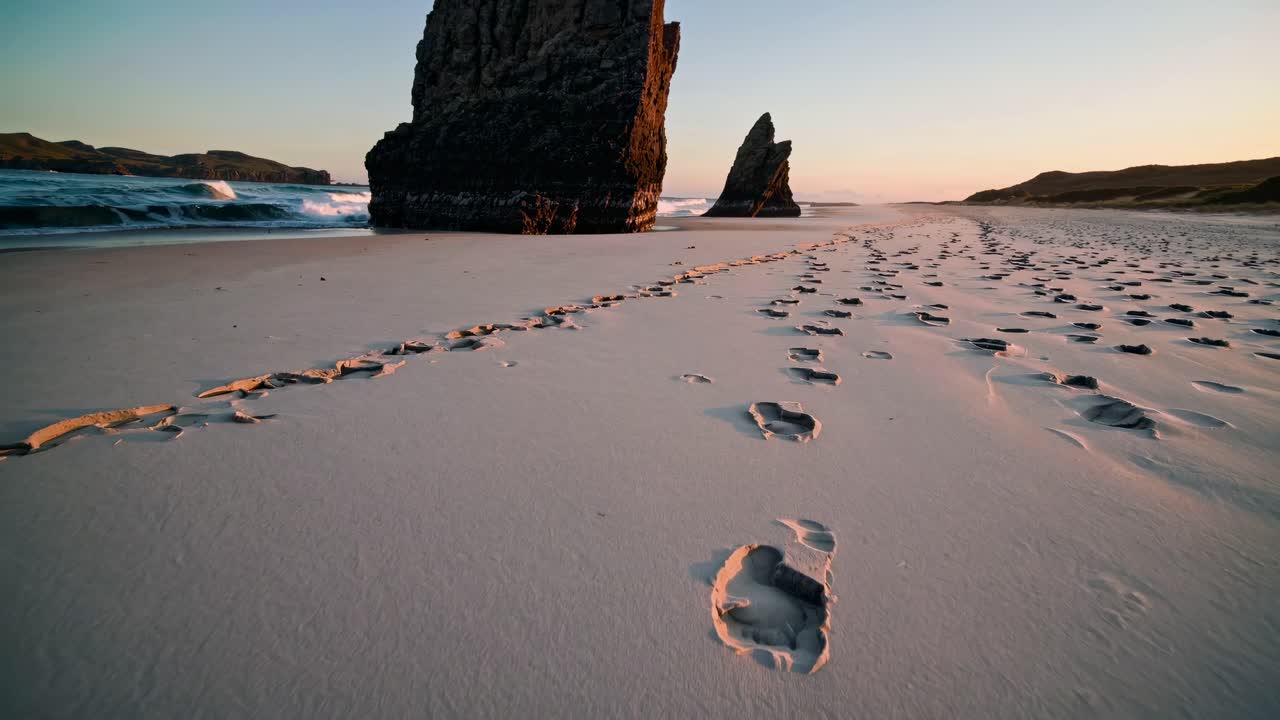 Low-angle video frame of a serene beach at sunset, capturing footprints leading to towering rock