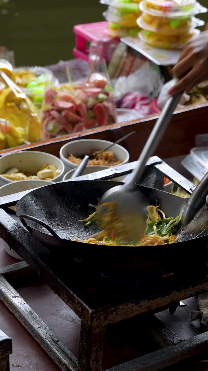 Penjual makanan jalanan menyiapkan mi Pad Thai tradisional di wajan di atas perahu di pasar terapung di Bangkok, Thailand. Close-up persiapan masakan Asia otentik di siang hari, vertikal