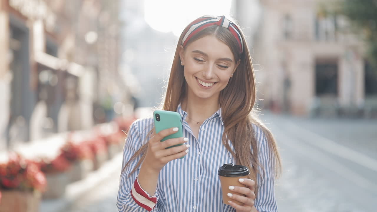 Giovane donna felice cammina con il caffè e usa lo smartphone in città.