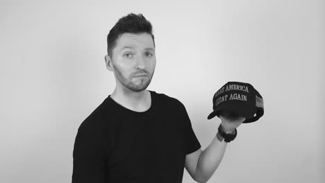 grayscale footage of tattooed man with short hair and beard holding up a black dark maga or make america great again hat with confidence and begins to nod and is nodding with approval and smiling.