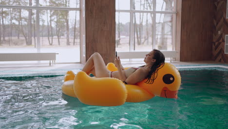 fin de semana en un baño termal mujer en bikini está flotando en un círculo inflable en la piscina usando un teléfono inteligente con internet