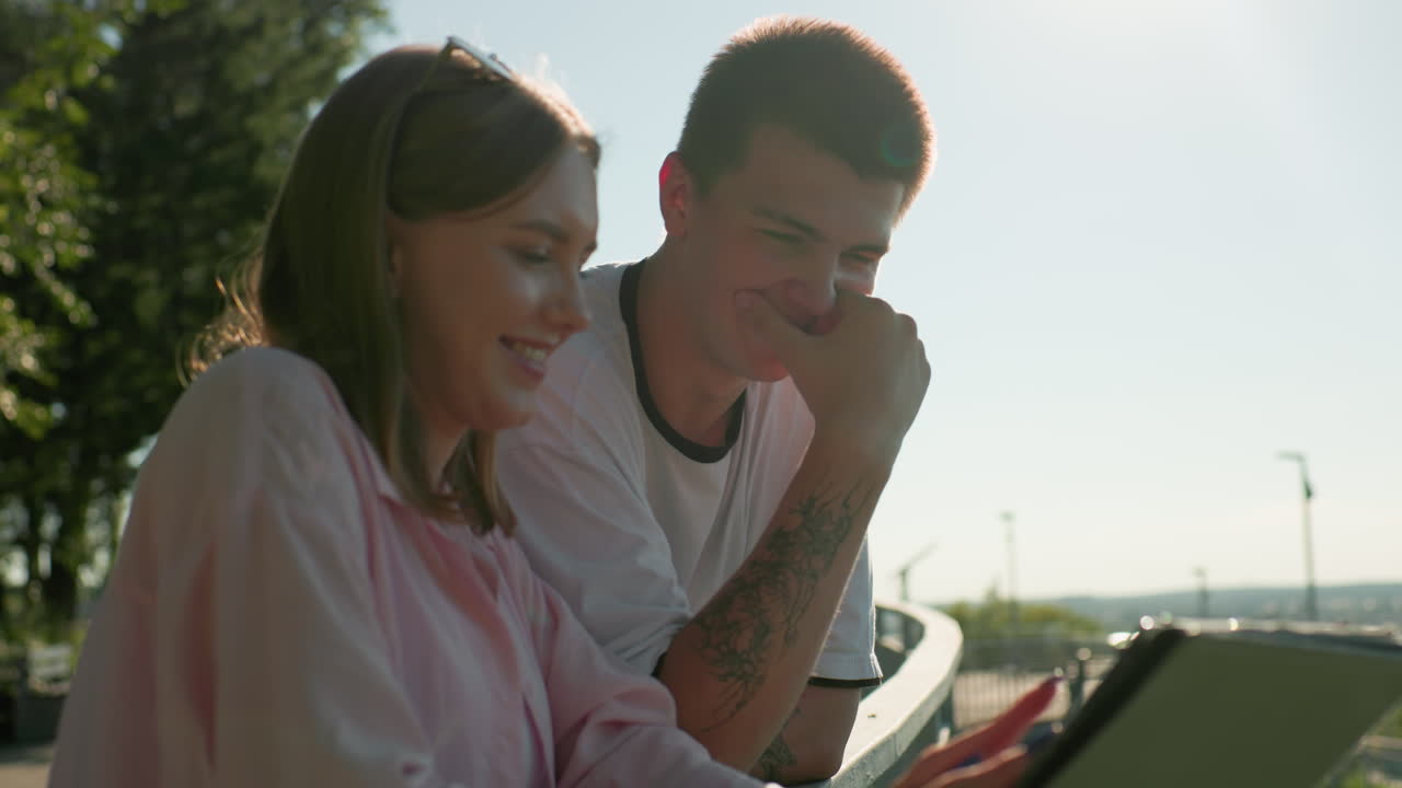 крупный план друзей, стоящих рядом, женщина показывает планшет парню, указывая на него, на заднем плане деревья, скамейка и расслабленная наружная обстановка, наполненная солнечным светом