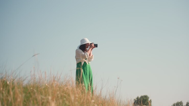 Woman in green dress and wide white hat walks through tall grass with camera in hand, enjoying peaceful exploration under clear sky while scouting and preparing to take landscape pictures