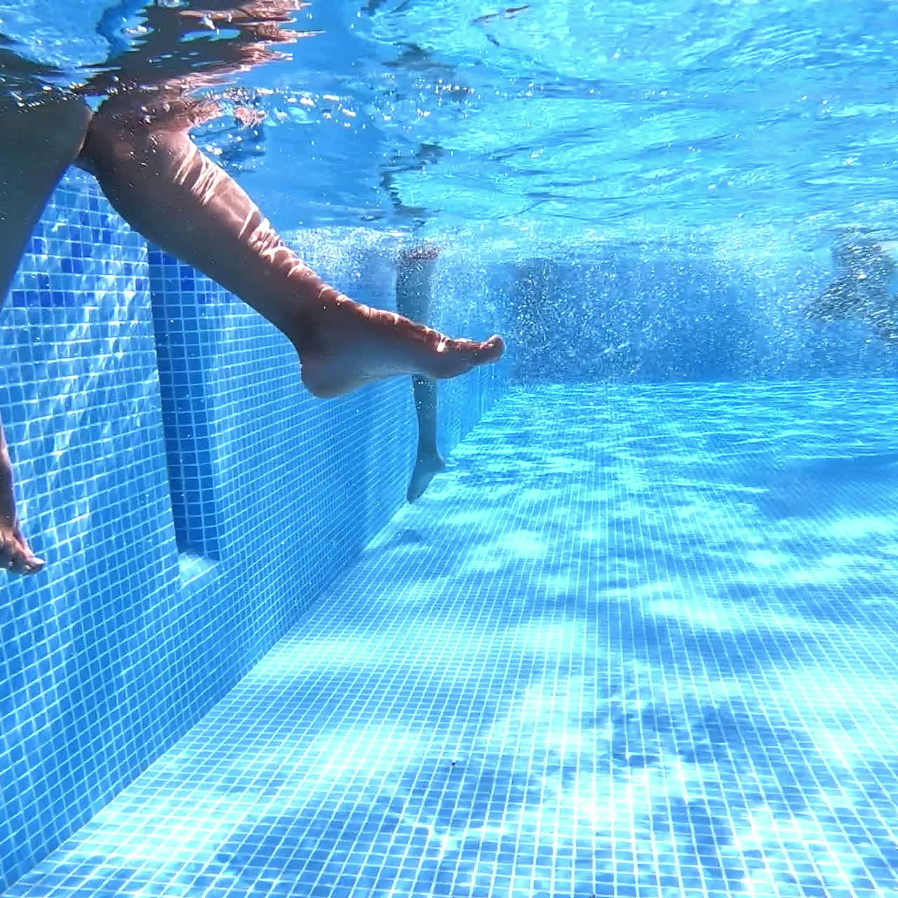 Female legs under water in the pool. Children are walking in the clear blue water of the swimming pool. Underwater camera shoot the video from the bottom of the pool.