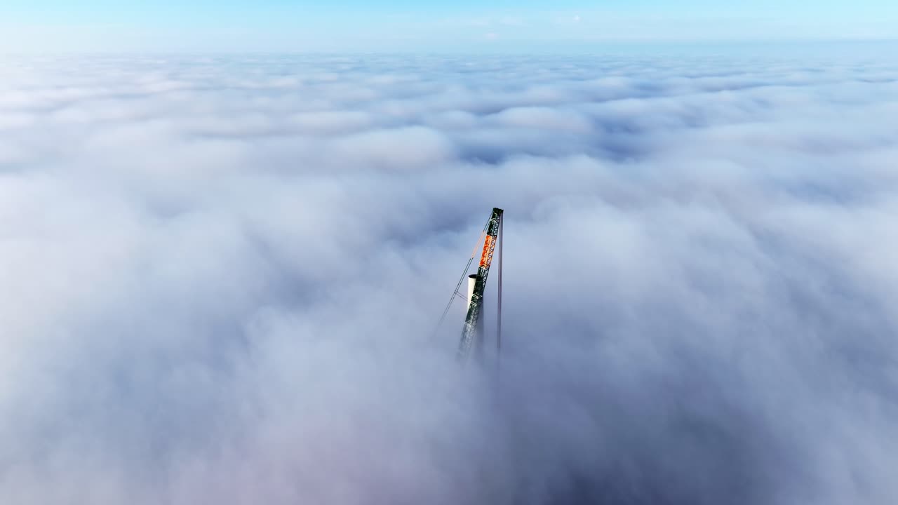 Impressive aerial view of a massive construction crane building the base of a wind turbine tower, with the top of the crane rising high above a thick layer of clouds in a Latvian wind farm at sunset