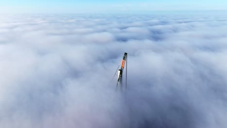 impressive aerial view of a massive construction crane building the base of a wind turbine tower, with the top of the crane rising high above a thick layer of clouds in a latvian wind farm at sunset