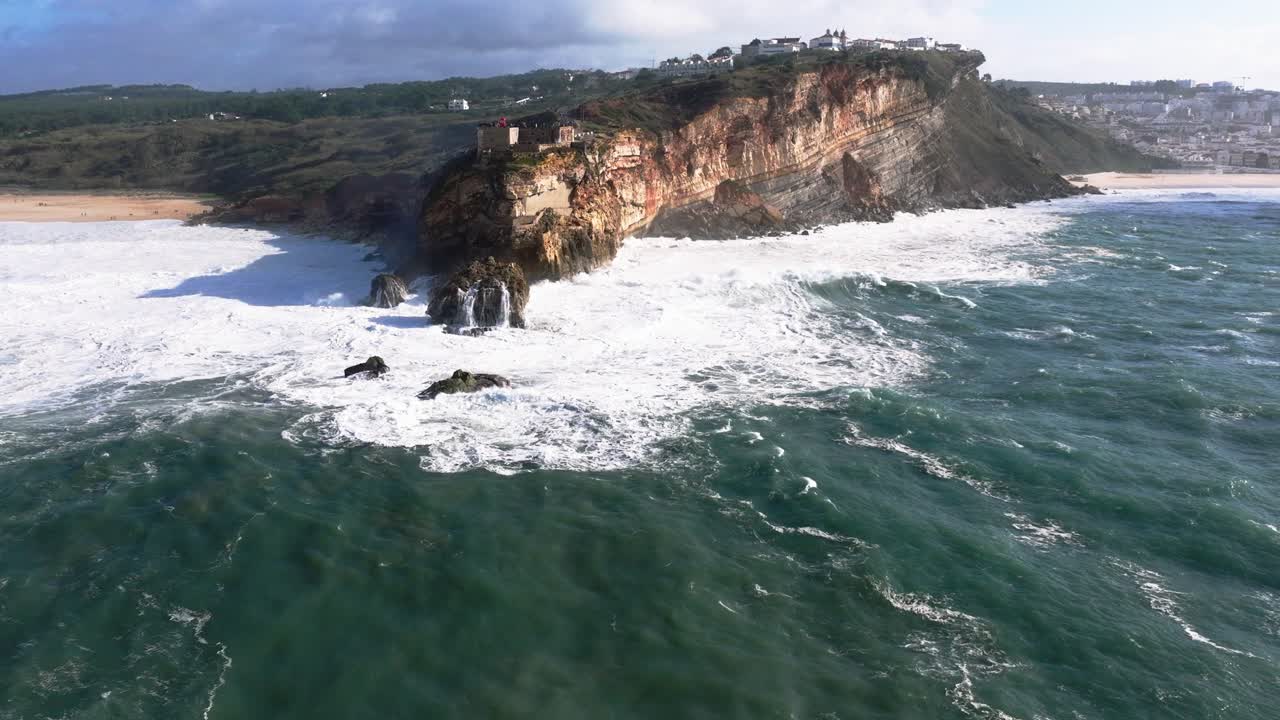 Nazaré, Portogallo, onde giganti che si infrangono sul promontorio roccioso. Vista aerea con drone del faro di Nazaré e delle turbine eoliche