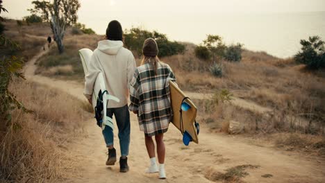 Rear-view-of-a-happy-couple,-a-brunette-guy-in-a-hat-in-a-white-sweatshirt-and-a-blonde-girl-in-a-hat-in-a-checkered-shirt-walk-down-the-slope-and-carry-boards-for-surfing-near-the-sea-in-the-summer