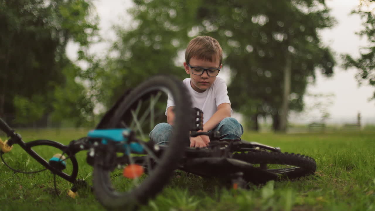 молодой мальчик приседает рядом со своим велосипедом, кажусь глубоко в мыслях, с размытием зеленых деревьев на заднем плане