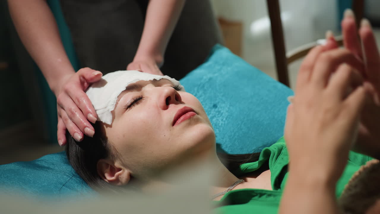 베개에 기대어 누워있는 아시아 여성, 간병인이 젖은 천을 이마와 눈썹에 올려놓고, 세심한 손길로 감은 눈을 진정시키고, 청록색 쿠션, 초록색 셔츠, 침대 옆 물병이 보이고, 저녁 조명, 차분함