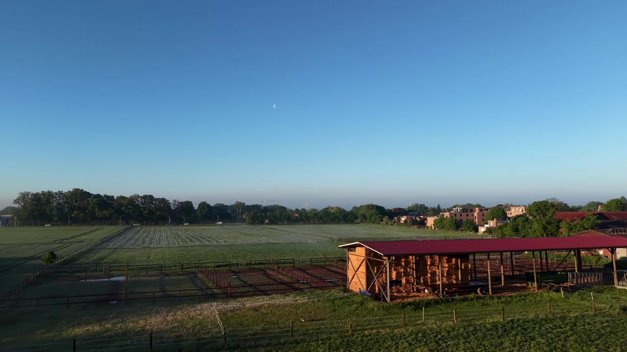 Paisagem rural agrícola com abrigo de feno de madeira, cercado para animais, vastos campos verdes abertos, telhados de vilarejos espalhados, linha densa de árvores e céu azul claro matinal ao nascer do sol baixo. Alemanha, Europa. Dolly aéreo