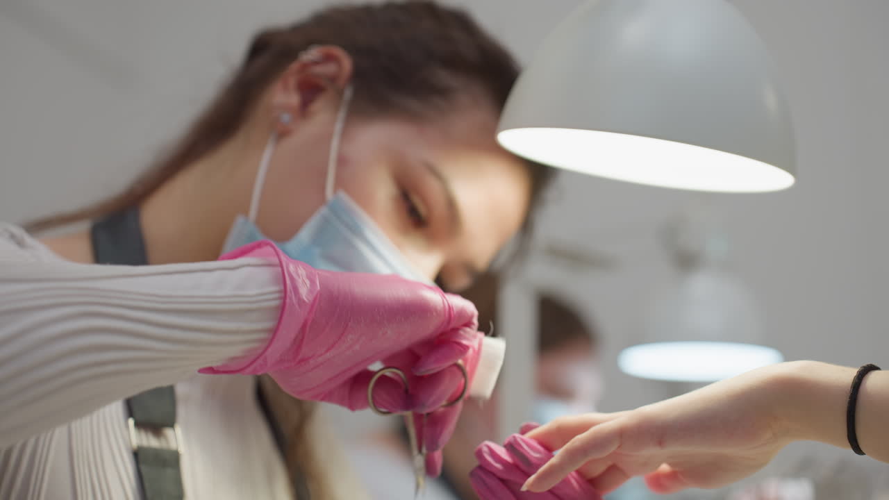 Closeup view of nail technician in mask and pink gloves trimming customer nails with scissors while customer hand rests on white platform, illuminated by bright overhead lamp in clean salon setting