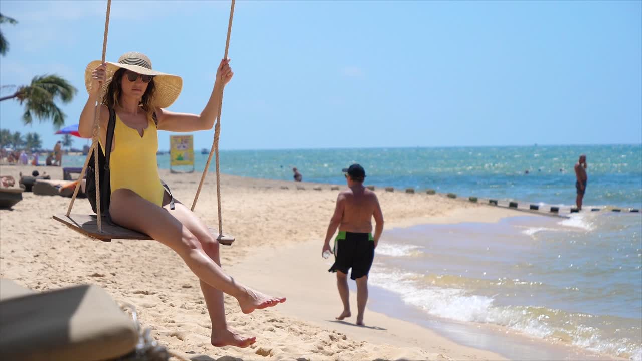 A young woman dressed in a swimsuit swings joyfully on a beach in Phu Quoc, Vietnam, while people enjoy the sun and water in the background, creating a lively atmosphere.