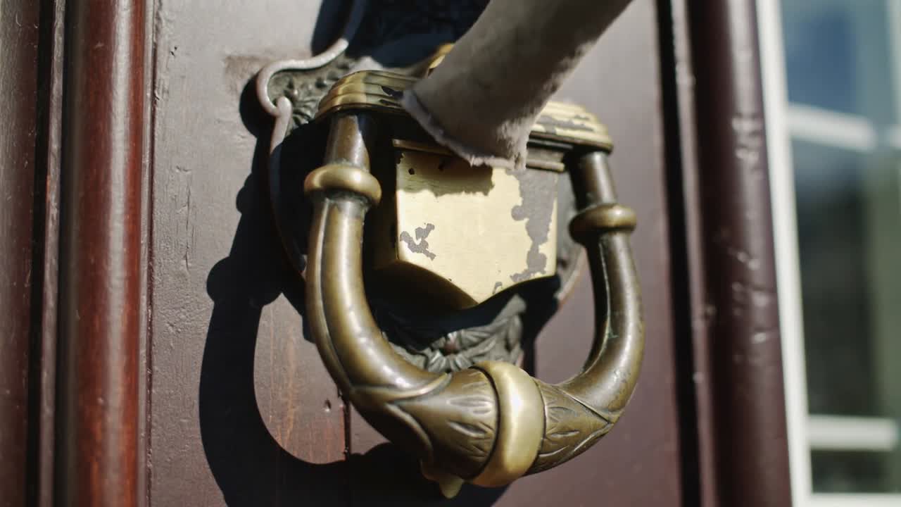 Braccio con manica grigia che entra, solleva un battente in ottone su una porta di legno scuro, con anello oscillante che segnala l'arrivo.