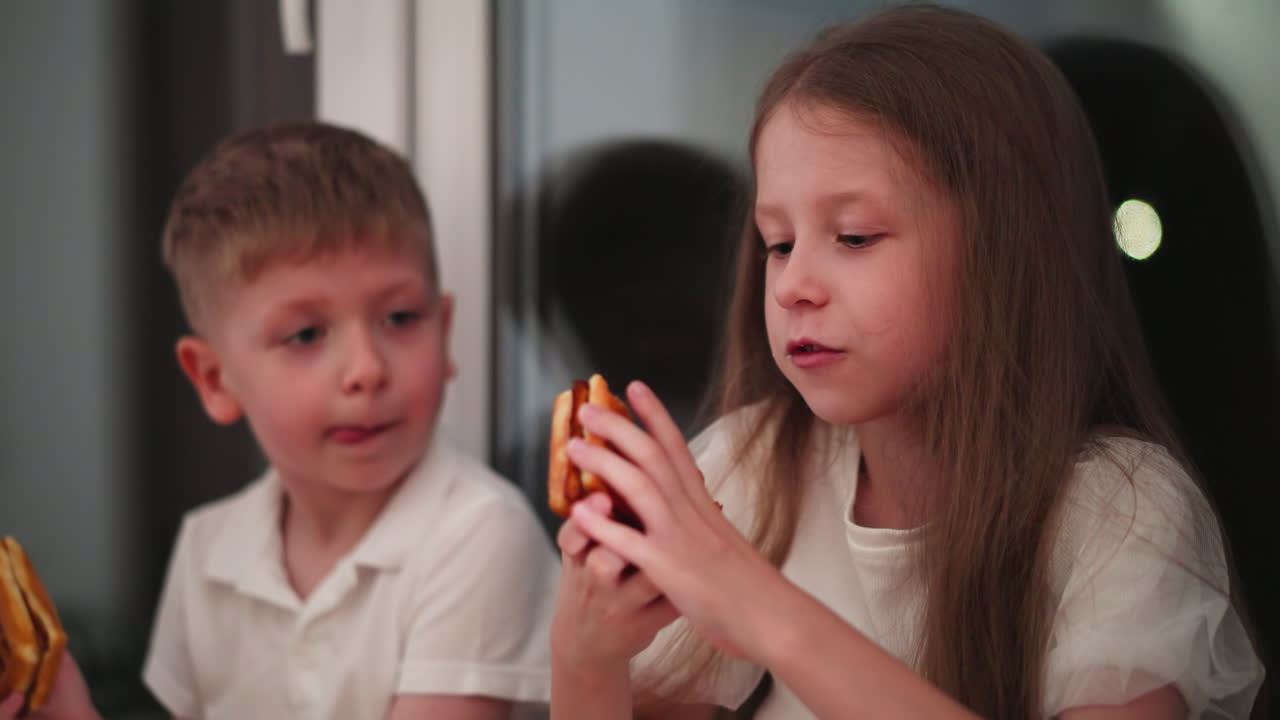 Children seated indoors beside glass door reflecting their image with visible bokeh lights outside, eating waffles and casually talking, relaxed moment during evening snack time