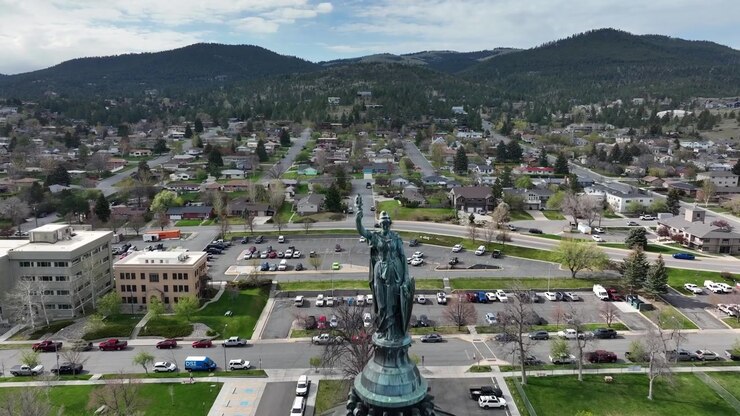 Sentiero circolare e aiuola a forma del Montana di fronte all'edificio del Campidoglio, vialetti curvi che incorniciano il prato sotto montagne boscose e cielo parzialmente nuvoloso. Helena, Montana, USA. Drone aereo in avvicinamento