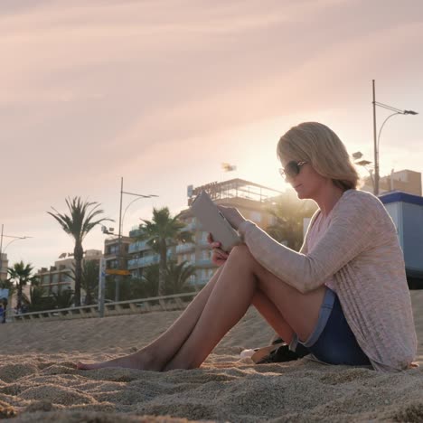 Eine-Junge-Frau-Benutzt-Ein-Tablet,-Das-Am-Strand-Vor-Dem-Hintergrund-Von-Palmen-In-Spanien-Sitzt